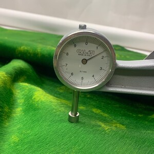 Może przedstawiać: Srebrny miernik tarczowy, oznaczony "CALATI MADE IN ITALY", mierzy grubość materiału barwionego na zielono i ż&oacute;łto. Miernik wskazuje około 2,0 na tarczy, co wskazuje na pomiar 0,2 mm. Materiał ma teksturowany wygląd, przypominający futro.