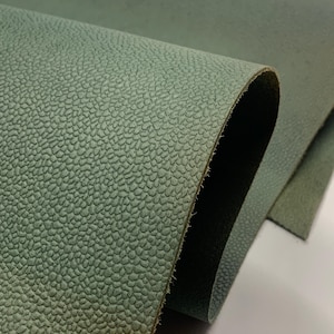 May include: Close-up of a sheet of sage green leather. The leather has a textured, pebble-like surface. The edges of the leather are visible, showing the layers and texture of the material. The leather is folded over.