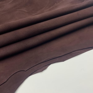 Könnte beinhalten: Nahaufnahme eines gefalteten Stücks dunkelbraunen Leders. Das Leder hat eine weiche, Wildleder-ähnliche Textur und eine satte, warme Farbe. Die Falten erzeugen Schatten und Lichtreflexe und verleihen dem Bild Tiefe. Das Leder scheint hochwertig und für verschiedene Bastelprojekte geeignet zu sein.
