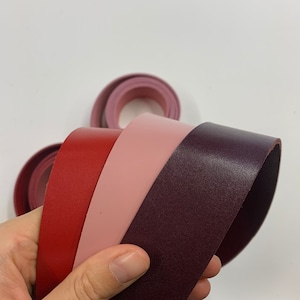 May include: Four leather straps in different shades of pink and burgundy. The straps are rolled up and held in a hand.