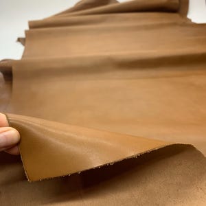 May include: A close-up of a sheet of tan-colored leather. The leather is folded over, revealing a smooth, finished surface. The edges of the leather are slightly rough. The leather appears to be supple and of high quality.