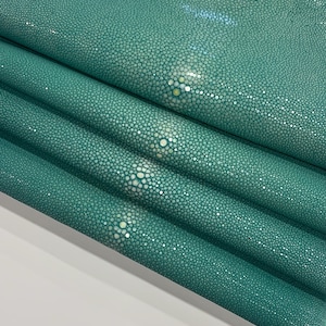 K&ouml;nnte beinhalten: Nahaufnahme von gefaltetem, teal-farbenem Rochenleder. Das Material hat eine strukturierte Oberfl&auml;che mit kleinen, eng gepackten, runden Erhebungen. Das nat&uuml;rliche Muster umfasst hellere, perlenartige Punkte, die eine einzigartige visuelle Textur erzeugen. F&uuml;r Luxusartikel.