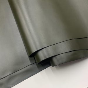May include: Close-up of olive green leather fabric, folded and layered. The material has a smooth, matte finish and a slight sheen. The edges of the leather are dark, creating a contrast. The fabric is likely used for crafting or upholstery.