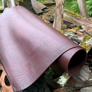 May include: A rolled sheet of burgundy leather. The leather has a textured surface and appears thick and durable. The roll is partially unraveled, revealing the inner layers. The background includes weathered wood and greenery.