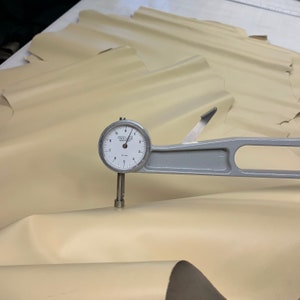 May include: A close-up of a leather thickness gauge measuring the thickness of a piece of beige leather. The gauge reads 0.1 mm.