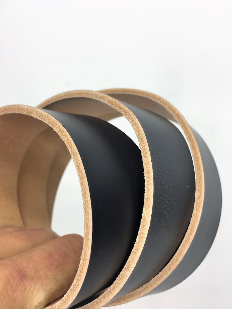 May include: Close-up of a rolled black leather belt with a natural tan edge. The leather is smooth and the edges are neatly finished. The belt is likely for use with trousers or skirts.