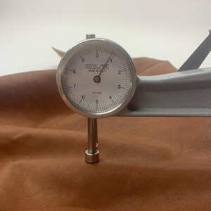 Puede incluir: Primer plano de un medidor de grosor de cuero que mide el grosor de un trozo de cuero marr&oacute;n. El medidor marca 0,1 mm.