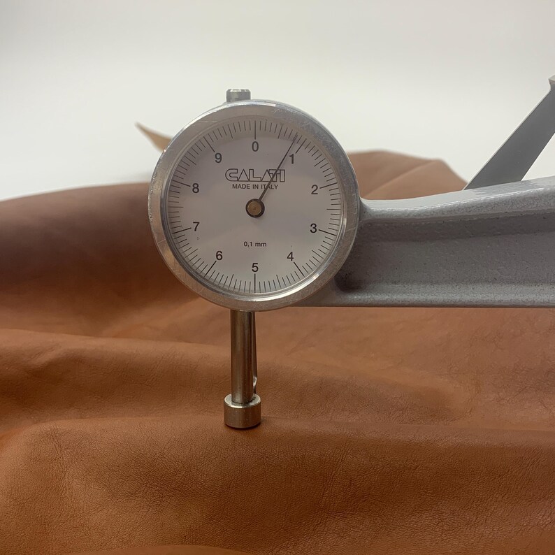 Puede incluir: Primer plano de un medidor de grosor de cuero que mide el grosor de un trozo de cuero marr&oacute;n. El medidor marca 0,1 mm.