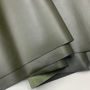 May include: Close-up of olive green leather sheets, folded and layered. The leather has a smooth, matte finish. The image highlights the texture and color variations of the material, suitable for crafting or upholstery projects.