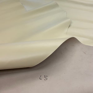 May include: A close-up of a cream-coloured fabric with a smooth, satin-like finish. The fabric is folded over itself, creating a soft, flowing drape. The fabric is marked with the number "L5".