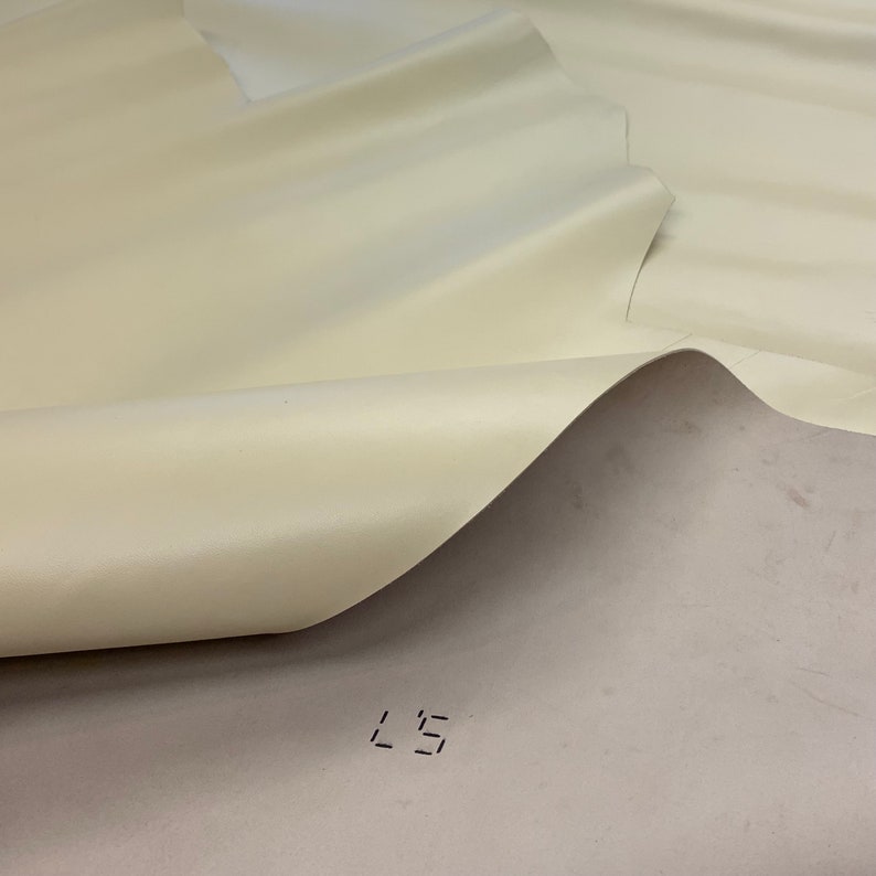 May include: A close-up of a cream-coloured fabric with a smooth, satin-like finish. The fabric is folded over itself, creating a soft, flowing drape. The fabric is marked with the number "L5".