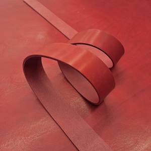 May include: A close-up of red leather strips, one straight and the other curled. The leather has a smooth, shiny surface and a deep red color. The background is a matching red leather surface.