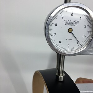 May include: A black leather strip is measured with a dial gauge. The gauge, labelled "CALATI MADE IN ITALY", shows a measurement of approximately 0.1 mm. The leather has a light tan underside.