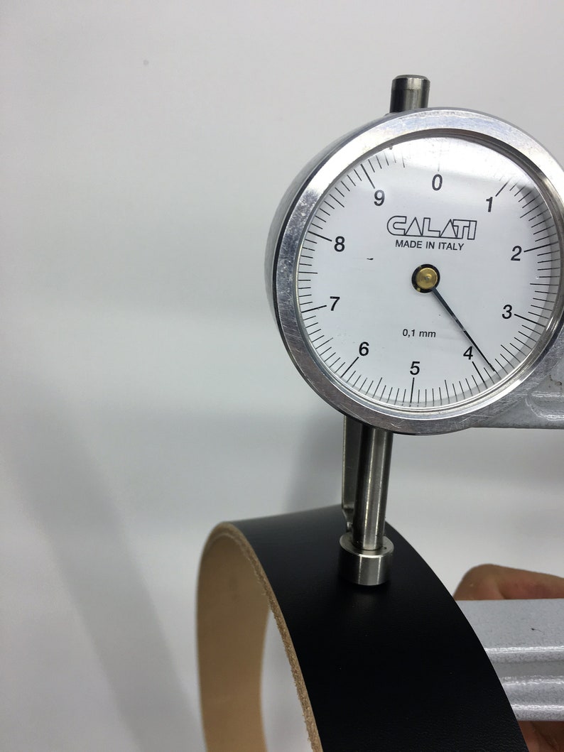 May include: A black leather strip is measured with a dial gauge. The gauge, labelled "CALATI MADE IN ITALY", shows a measurement of approximately 0.1 mm. The leather has a light tan underside.