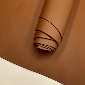 May include: A roll of smooth, brown leather. The leather is a warm, earthy tone, with a slight sheen. The edges of the roll are visible, showing the thickness of the material. The leather is likely for crafting or upholstery.
