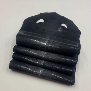 May include: Folded black stingray leather pieces. The leather has a textured surface with small, light-coloured dots. The top piece has two circular cutouts. The edges of the leather are visible, showing the layers.