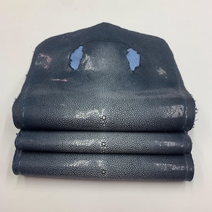Op de afbeelding: Een stapel van drie gevouwen stukken marineblauw roggenleer. Het leer heeft een textuur met kleine, witte, parelachtige stippen. Het bovenste stuk heeft twee onregelmatige blauwe vlekken.