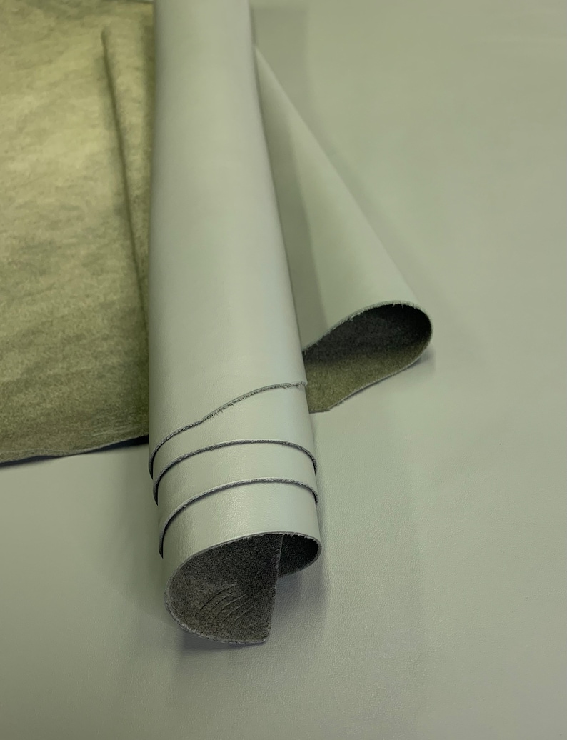 May include: A roll of light gray leather with a dark gray backing. The leather is rolled up and the backing is visible on the inside of the roll.