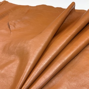 Può includere: Primo piano di un foglio di pelle color marrone chiaro. La pelle è piegata, mostrando la sua consistenza liscia e le variazioni di colore. Il materiale appare morbido e pronto per la lavorazione.