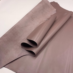 May include: Two sheets of mauve-colored leather are displayed, one partially rolled. The leather has a smooth, shiny finish, with a slight sheen. The other sheet has a textured, matte appearance. The sheets are arranged on a white surface.