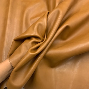 May include: A close-up shot of a sheet of tan leather, showing folds and creases. The leather has a smooth, matte finish and a warm, golden-brown colour. The material appears soft and pliable, suggesting it is suitable for various crafting projects.