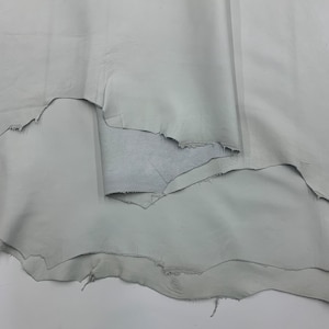 Pode incluir: Várias peças de couro cinza claro são sobrepostas. As bordas são irregulares e rasgadas, revelando a textura do material. O couro tem um acabamento liso e fosco. Ideal para artesanato e marroquinaria.