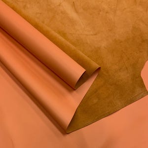 Puede incluir: Primer plano de hojas de cuero naranja, una enrollada y la otra plana. La hoja enrollada revela un tono más claro de naranja, mientras que la hoja plana muestra una superficie texturizada más oscura. El fondo presenta una hoja de cuero marrón.