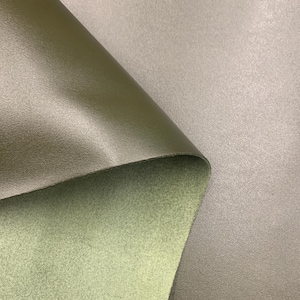 May include: Close-up of olive green and gray leather fabric. The top layer is smooth and shiny, while the underside is a textured, lighter green. The gray fabric has a subtle, speckled appearance. The layers are folded over each other.