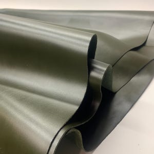 May include: Close-up of olive green leather fabric, showing the smooth texture and folds of the material. The lighting highlights the sheen of the leather, suggesting a high-quality product. The fabric is likely used for crafting or upholstery.