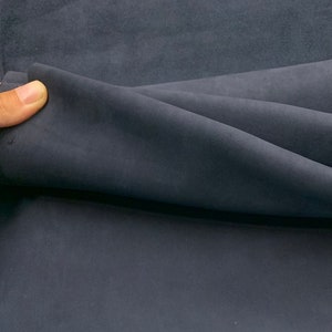 May include: A piece of dark navy blue fabric with a soft, matte, suede-like texture. The material is folded over to show its flexible drape and consistent deep blue color across the surface, highlighting the smooth, non-reflective finish.