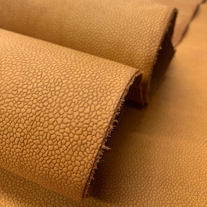 May include: Close-up of tan leather with a textured, pebble-like pattern. The leather is folded, revealing the rough, unfinished edge. The material has a warm, earthy tone, suggesting it could be used for various crafting projects.
