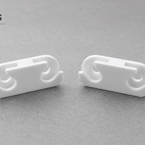 May include: Two white plastic clips with a symmetrical, elongated shape featuring two circular cutouts and a central bridge. The text in3dlabs INTERIOR DESIGN is visible in the top left corner, and a German flag icon is in the top right corner.