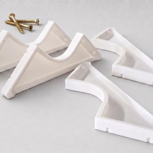 May include: A set of four white plastic corner shelf brackets with curved cutouts and two gold-colored metal screws. The brackets feature a triangular shape with a recessed center section and flat edges designed for mounting into corners.