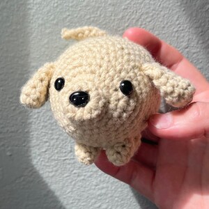 May include: A crocheted dog toy in a light beige color. The dog has black eyes and a black nose. The ears and legs are small and rounded. The toy is handmade and has a textured appearance.