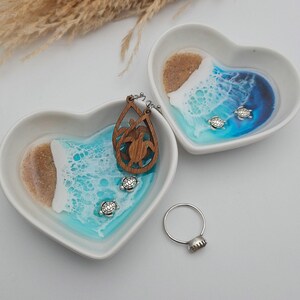 May include: Two white heart-shaped ceramic dishes featuring a resin ocean design with sand, turquoise and blue water, and white foam. The dishes contain wooden teardrop-shaped turtle earrings, small silver turtle charms, and a silver ring.