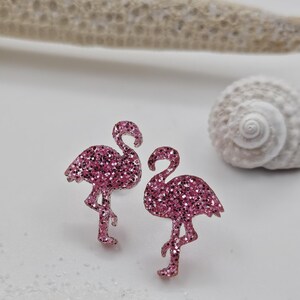 K&ouml;nnte beinhalten: Ein Paar Ohrstecker in Form von Flamingos mit einer leuchtend pinken Glitzerbeschichtung. Jeder Flamingo ist auf einem Bein stehend mit nach unten gebogenem Hals dargestellt und weist eine texturierte, funkelnde Oberfl&auml;che auf.