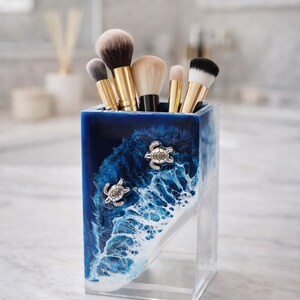 May include: A rectangular resin makeup brush holder featuring a deep blue and white ocean wave design with two silver-toned sea turtle embellishments. The container is filled with five makeup brushes that have gold-toned handles and bristles.