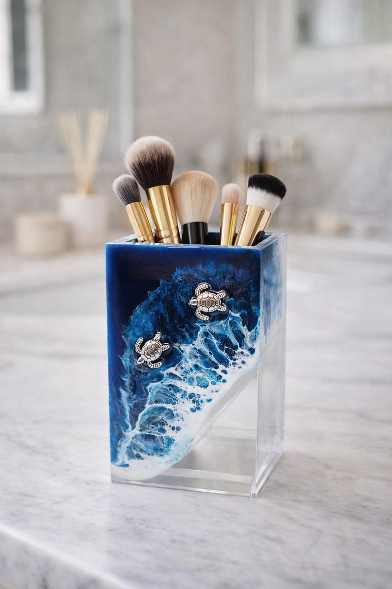 May include: A rectangular resin makeup brush holder featuring a deep blue and white ocean wave design with two silver-toned sea turtle embellishments. The container is filled with five makeup brushes that have gold-toned handles and bristles.