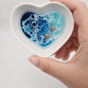 May include: A heart-shaped white ceramic dish featuring a resin inlay that mimics ocean waves in shades of deep blue, turquoise, and white foam. Two small, metallic silver-toned sea turtle charms are embedded within the resin surface.
