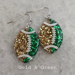 Op de afbeelding: Gouden en groene glitter voetbal oorbellen. De oorbellen zijn gevormd als een voetbal en hebben een witte omtrek. De voetbal is in twee&euml;n gedeeld, met een helft gouden glitter en de andere helft groene glitter.