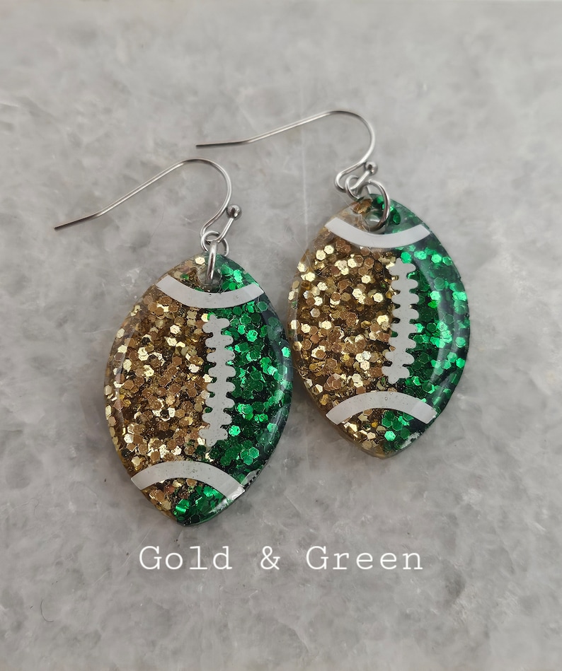 Op de afbeelding: Gouden en groene glitter voetbal oorbellen. De oorbellen zijn gevormd als een voetbal en hebben een witte omtrek. De voetbal is in twee&euml;n gedeeld, met een helft gouden glitter en de andere helft groene glitter.