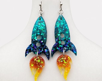 May include: A pair of dangle earrings shaped like rockets. The rocket bodies are made of teal and dark blue resin with embedded glitter. Attached to the base of each rocket is a flame-shaped charm in orange and yellow resin with glitter.