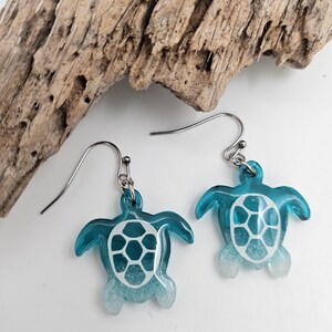 May include: A pair of dangle earrings featuring sea turtle charms made of translucent blue and white resin. The turtle shells have a white geometric pattern, and the earrings are attached to silver-toned metal hooks for pierced ears.