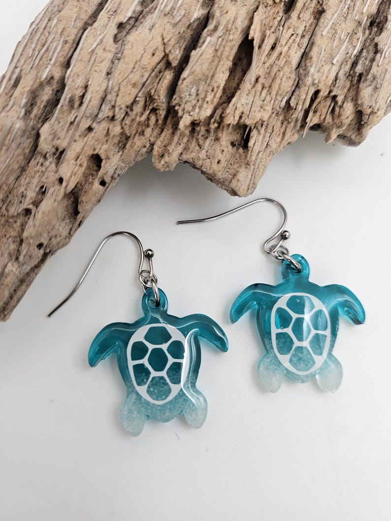 May include: A pair of dangle earrings featuring sea turtle charms made of translucent blue and white resin. The turtle shells have a white geometric pattern, and the earrings are attached to silver-toned metal hooks for pierced ears.
