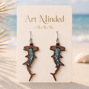 May include: A pair of hammerhead shark earrings made of dark brown wood with a central inlay of blue resin filled with iridescent glitter. The earrings are attached to a cream-colored display card featuring the text Art Minded in gold font.