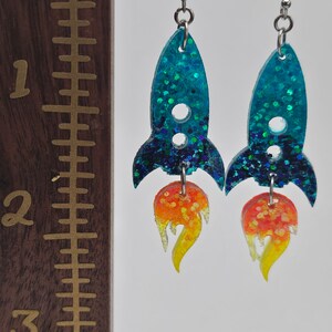 May include: A pair of handmade earrings shaped like rockets. The rocket bodies are teal with glitter, and the flames are orange and yellow. The earrings are approximately 2 inches long.