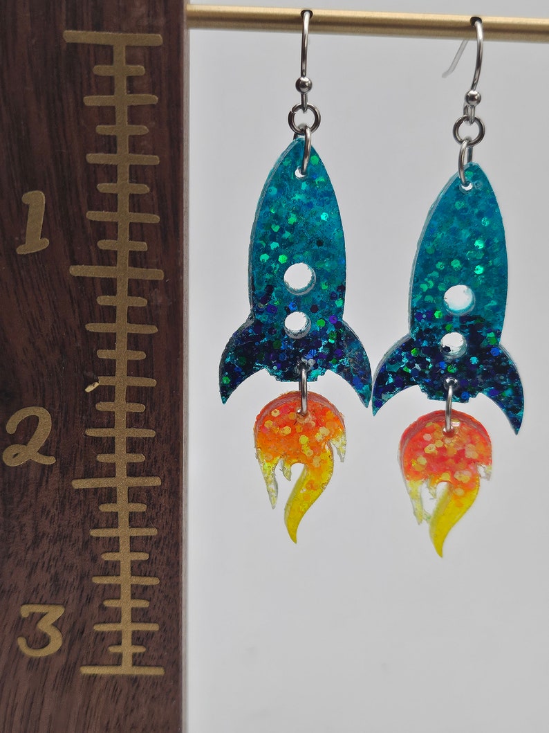 May include: A pair of handmade earrings shaped like rockets. The rocket bodies are teal with glitter, and the flames are orange and yellow. The earrings are approximately 2 inches long.