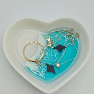 May include: Heart-shaped trinket dish with a turquoise and white resin design, featuring a gold ring and a delicate gold necklace with a small diamond pendant. The dish also has two black manta ray silhouettes.