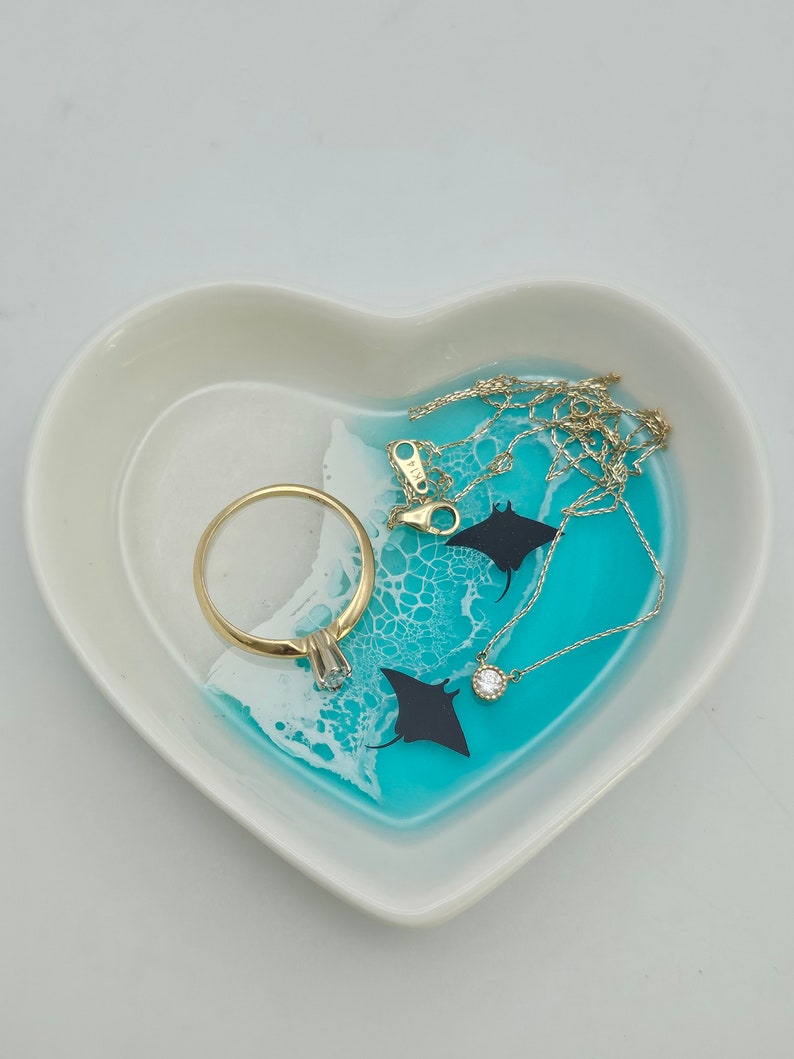 May include: Heart-shaped trinket dish with a turquoise and white resin design, featuring a gold ring and a delicate gold necklace with a small diamond pendant. The dish also has two black manta ray silhouettes.