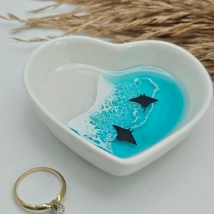 May include: A white heart-shaped trinket dish with a blue and white ocean-themed design. Two black manta ray silhouettes swim in the turquoise water. A gold ring with a diamond sits next to the dish.